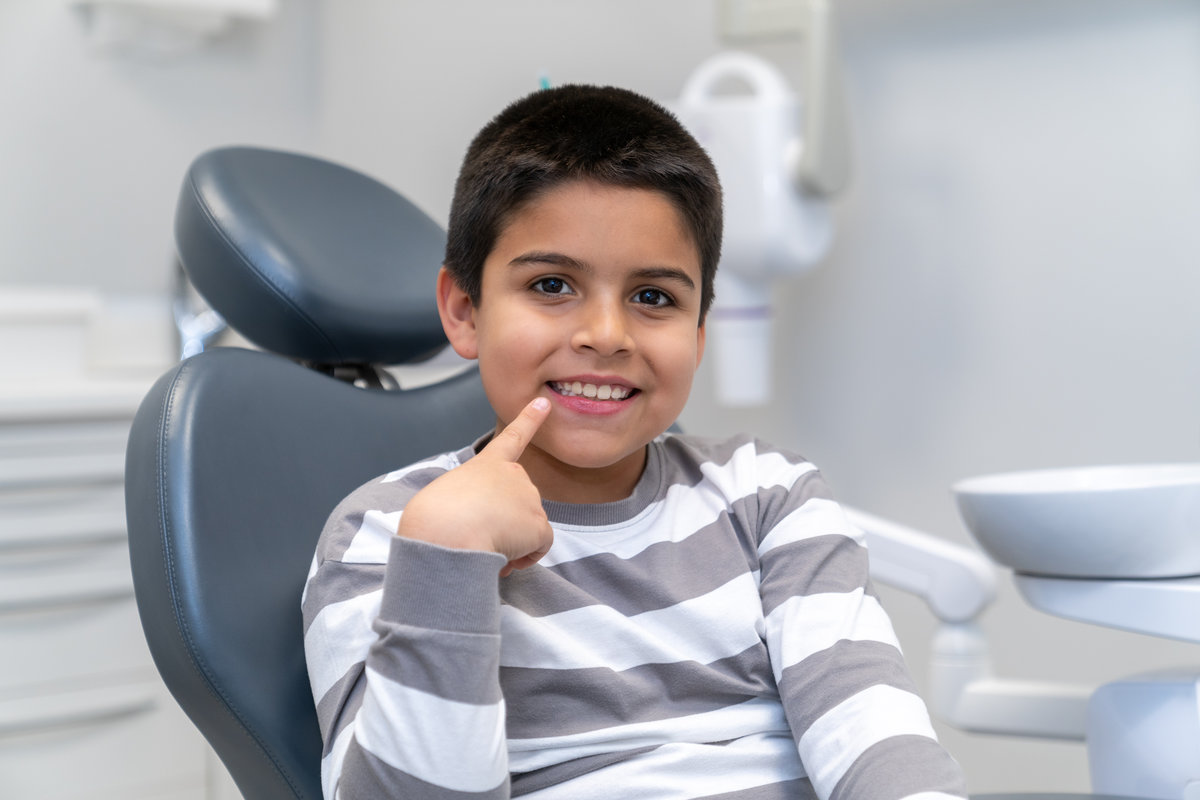 Smiling boy pointing his finger at his healthy teeth while sitting in dentist chair at clinic