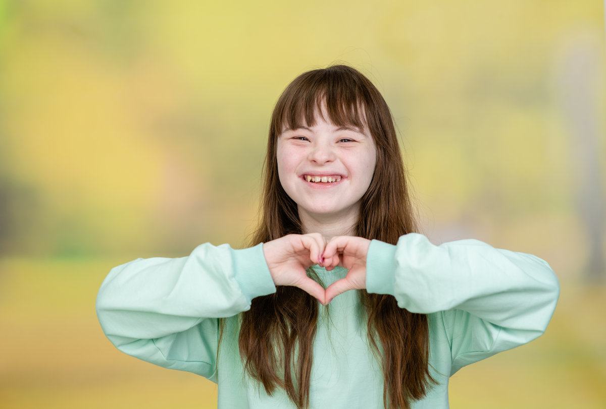 Happy young girl with Downs syndrom shows heart sign at summer park.