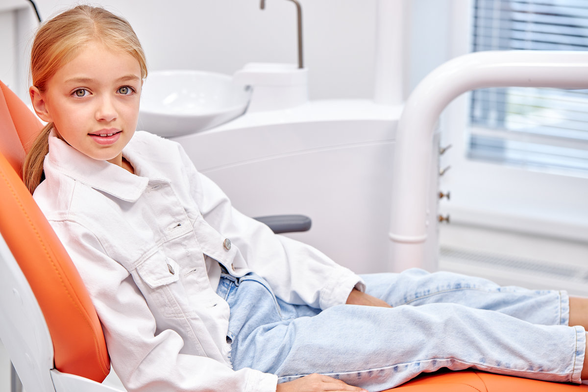Portrait of attractive little girl sitting on dental couch looking at camera, waiting for teeth treatment. Caucasian child in casual outfit in modern bright clinic. healthcare, medicine concept