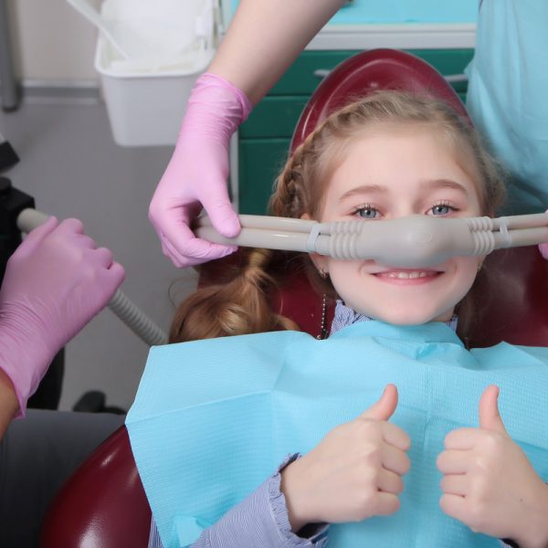 A little girl is comfortable to treat her teeth under superficial sedation. The girl smiles and holds two thumbs up. Milk teeth treatment. Treatment of children's teeth with nitrous oxide.