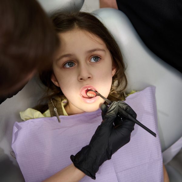 Close-up Caucasian little child girl with open mouth, is being examined by a dental hygienist, dentist doctor in pediatric dentistry clinic. Dental light directed at her oral cavity. Dental practice